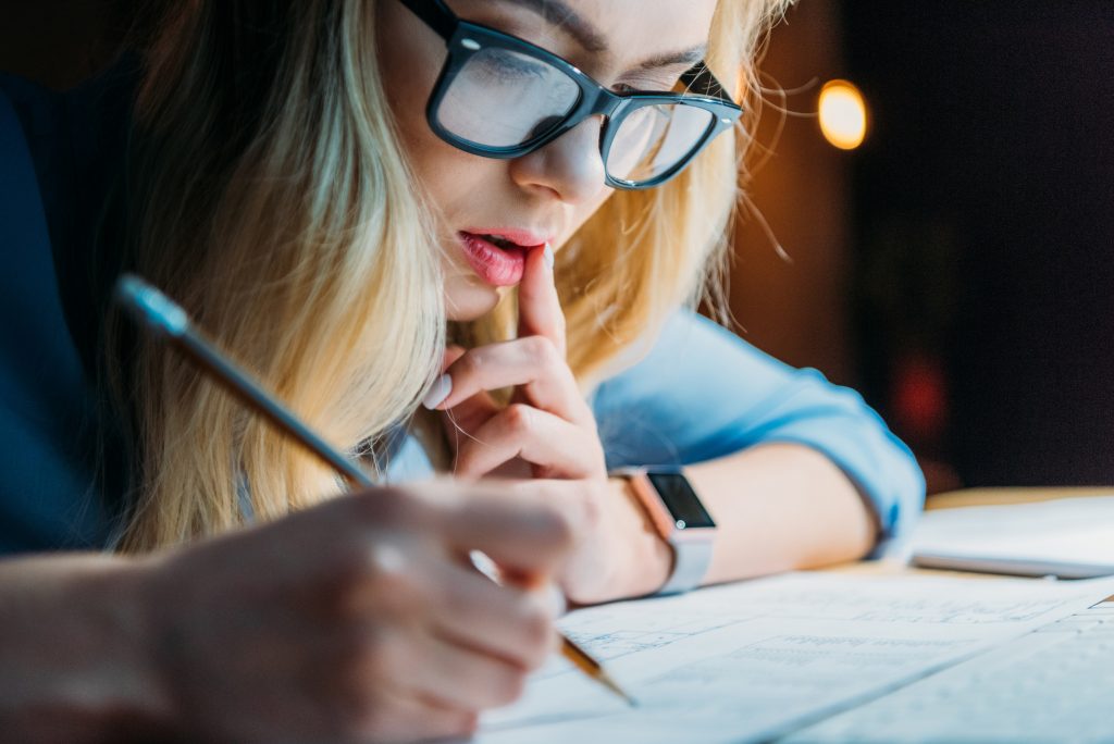 young blonde caucasian student in eyeglasses thinking and writing something with pencil, studying till late